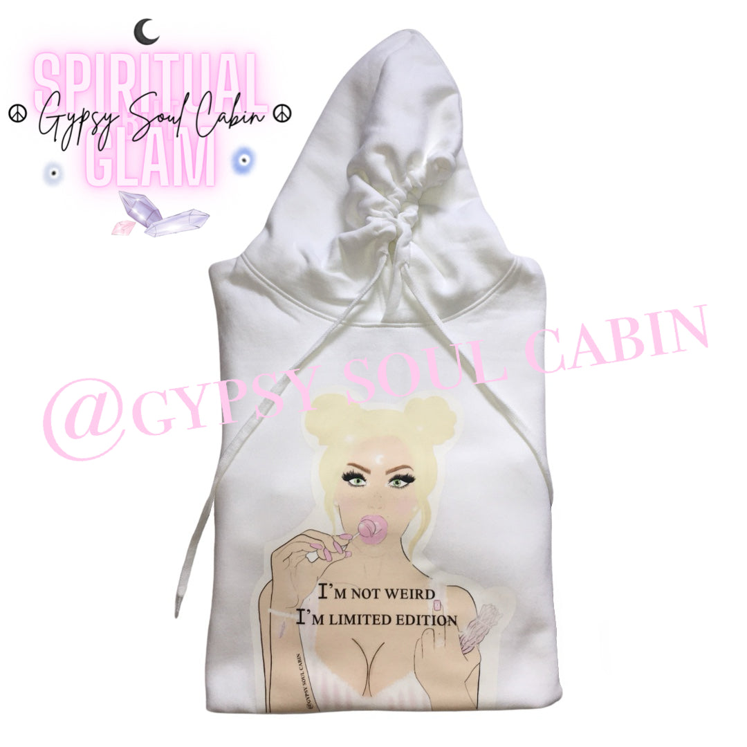 A white oversized hoodies with a graphic of a woman with blonde hair, holding a lollipop, and the text "I'M NOT WEIRD, I'M LIMITED EDITION" printed on the front. The design features pastel colours and a playful, artistic style.