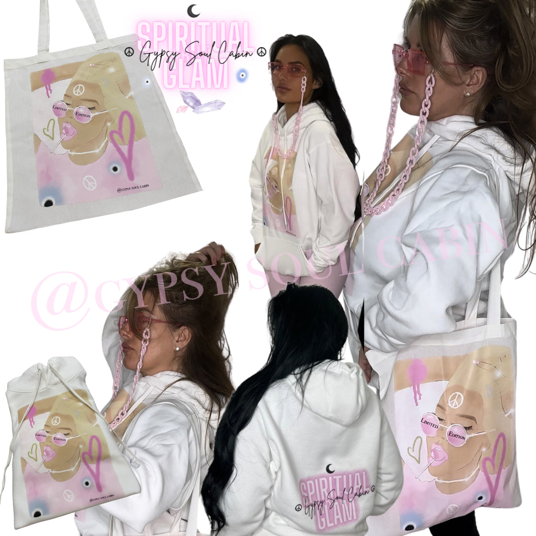 Group of women wearing white oversized hoodies featuring colorful spiritual girl artwork, with one holding a matching tote bag that displays the same art, pink necklaces, and the back of one hoodie showing "Spiritual Glam" text and a crescent moon.