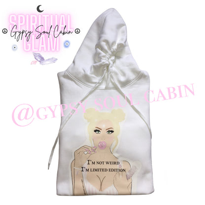 A white oversized hoodies with a graphic of a woman with blonde hair, holding a lollipop, and the text "I'M NOT WEIRD, I'M LIMITED EDITION" printed on the front. The design features pastel colours and a playful, artistic style.