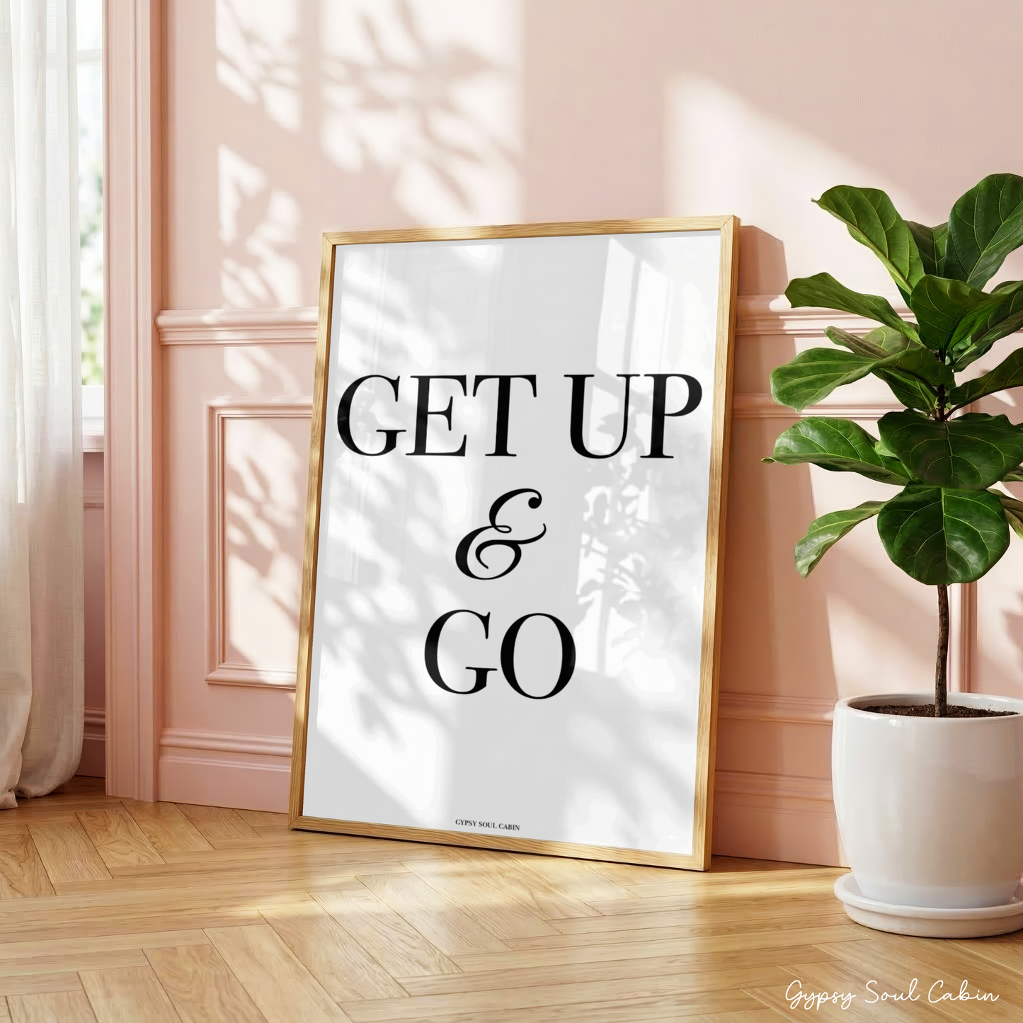 Minimalist black and white manifestation affirmation typography print reading Get Up & Go with a cursive ampersand in a large oak frame leaning against a panelled pink wall.