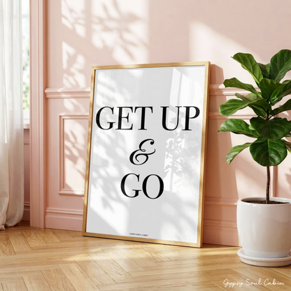 Minimalist black and white manifestation affirmation typography print reading Get Up & Go with a cursive ampersand in a large oak frame leaning against a panelled pink wall.