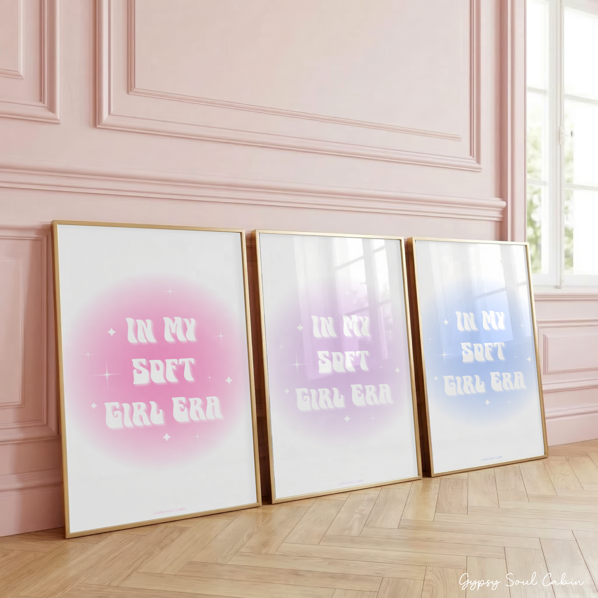 Gold-framed art prints leaning against a pink wall showcasing three colour options: pink, purple, and blue aura gradient glows with retro wavy text reading In My Soft Girl Era.