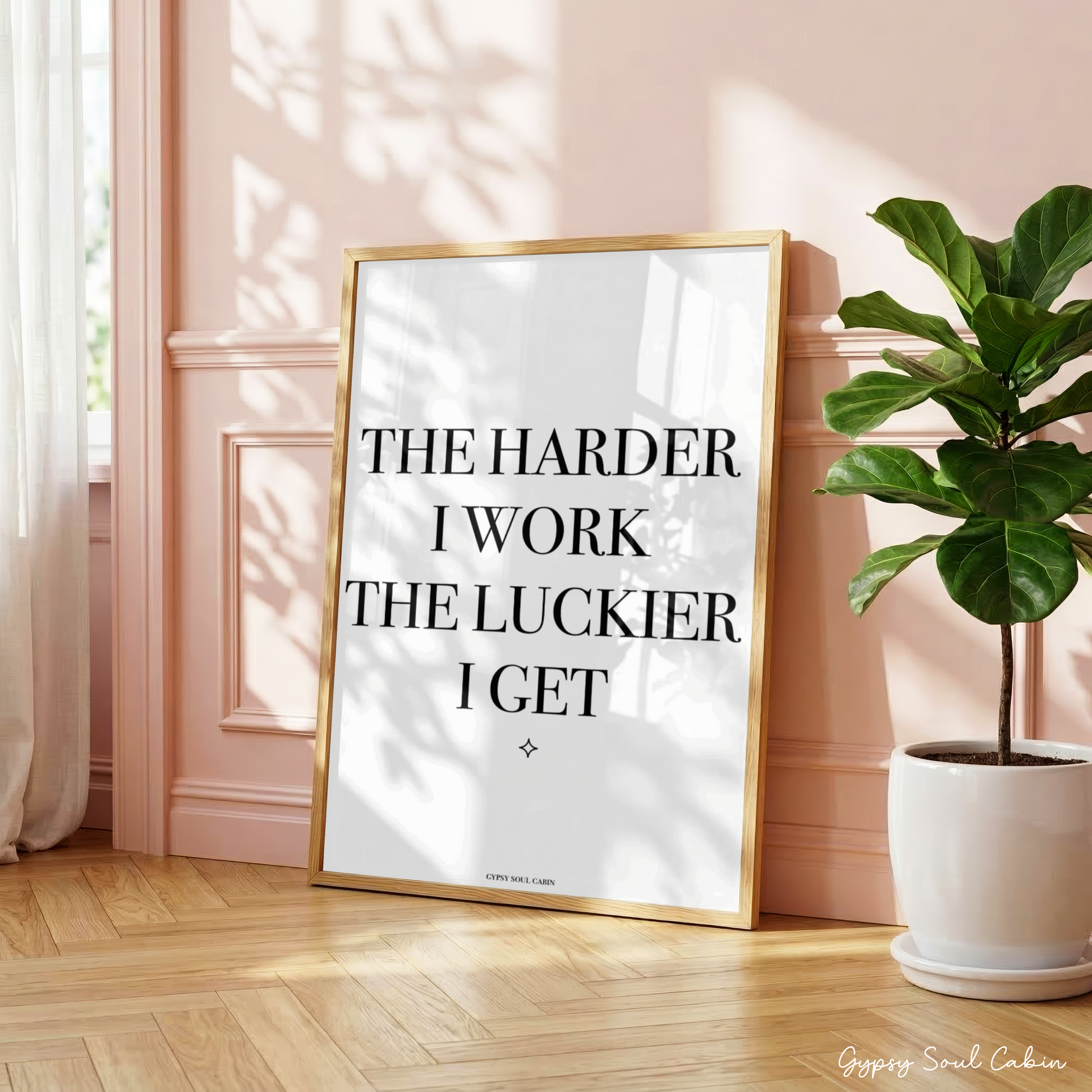 Minimalist black and white manifestation affirmation typography print reading The Harder I Work The Luckier I Get with a tiny star accent in a large oak frame leaning against a panelled pink wall.